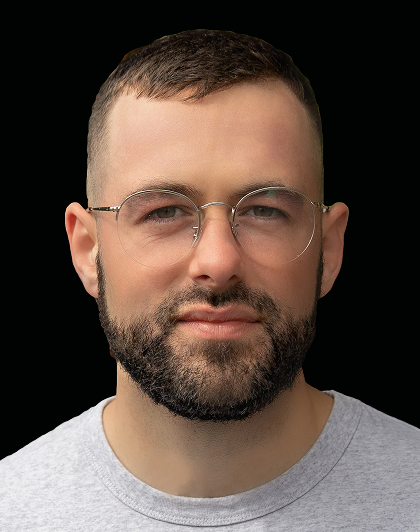 Portrait d'un homme aux cheveux bruns courts, portant des lunettes rondes et une barbe, regardant l'objectif devant un fond noir uni.