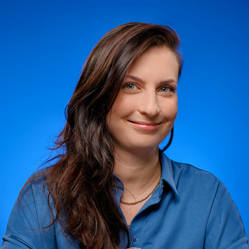 Portrait of a smiling woman with long brown hair wearing a blue button-down shirt against a blue backdrop.