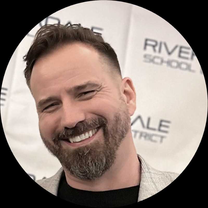 Smiling man with short dark hair and a beard in a circular portrait, background shows event signage.