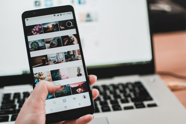 a person holding a mobile phone with open instagram account, laptop in the background