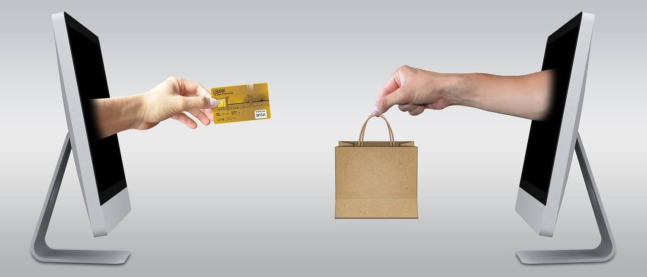 two hands emerging from computer screens, one holding a paper bag the other a credit card