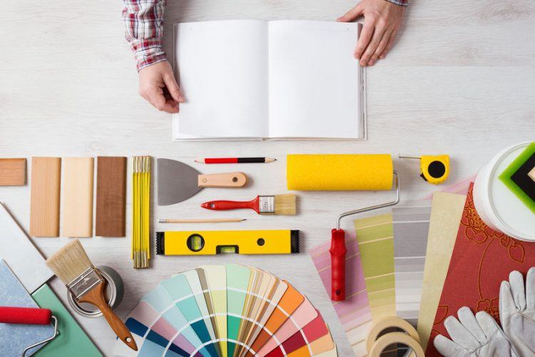 Hands holding DIY training open manual with work tools, color swatches and painting rollers at bottom, top view